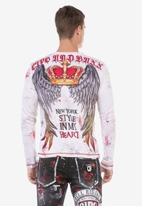 Langärmliges weißes Hemd mit roten Flecken, das eine Krone, Engelshügel und Text zeigt. Getragen mit schwarzen Jeans, die mit grafischen Details verziert sind.