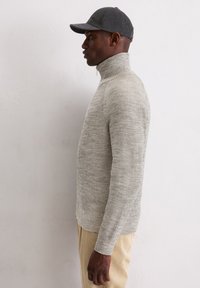 Lichtgrijze geribbelde gebreide pullover met een hoge kraag. Het model draagt een grijze pet en beige corduroypantalon, zijwaarts weergegeven tegen een neutrale achtergrond.