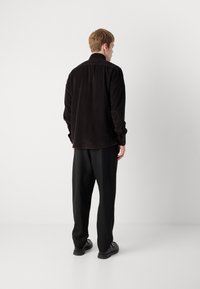Black corduroy shirt with a collared neck, long sleeves, and buttoned cuffs, paired with loose black trousers and dark shoes.