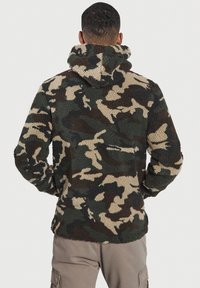 Sudadera de forro polar con patrón de camuflaje en verde, marrón y beige. Presenta una textura suave, un cuello alto y una capucha amplia.