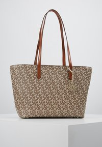 Sac fourre-tout en tissu beige avec un motif géométrique marron clair, équipé de deux anses en cuir marron et d'un charm décoratif attaché.