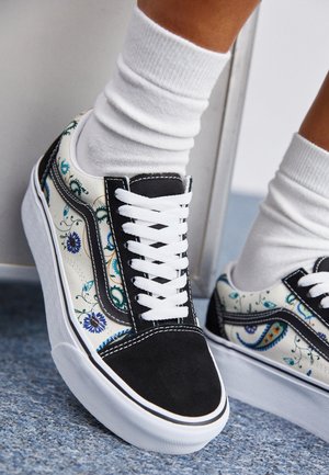 Persona que lleva zapatillas negras y blancas con estampado floral, cordones blancos y calcetines blancos, de pie sobre una alfombra azul.