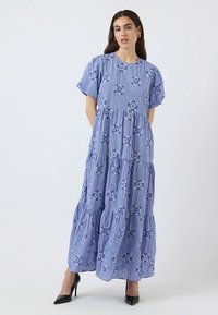 Mėlyna pledinė maxi suknelė su trumpomis pūstomis rankovėmis, sluoksniuotu dizainu, gėlėtu siuvinėjimu ir lygiu medvilnės audiniu. Juodi aukšti kulniukai.