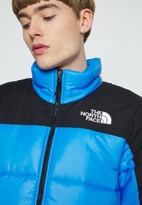 Blå og sort pufferjakke med høj krave, lynlås lukning og broderet logo. Har en skinnende overflade og quiltet design.