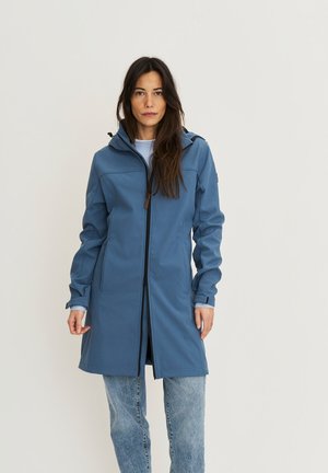 Femme aux longs cheveux foncés portant un manteau à capuche bleu sur un jean, debout devant un fond clair uni, face à l'avant.