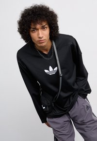 Sort langærmet mesh skjorte med et hvidt Adidas-logo, parret med grå bukser og en lille sort crossbody taske med et logo.