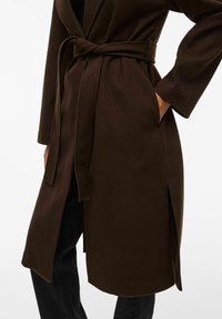 Manteau en mélange de laine marron avec une taille ceinturée, des manches longues et des poches latérales. Il présente une coupe droite et une fente à l'ourlet pour plus de flexibilité.