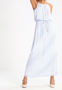 Donna che indossa un abito maxi senza maniche, con una fantasia blu e bianca, vita stretta e scarpe con tacco alto bianche, mentre posa con un braccio alzato.