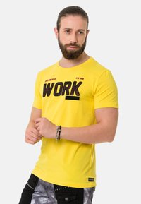 Gelbes Baumwoll-T-Shirt mit einem auffälligen schwarzen "WORK"-Grafik. Rundhalsausschnitt und kurze Ärmel. Getragen mit schwarzen gemusterten Hosen und Armbändern.
