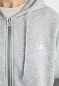 Felpa grigia chiara con zip in tessuto morbido e testurizzato. Presenta una zip frontale, cordini e un piccolo logo bianco sul petto.