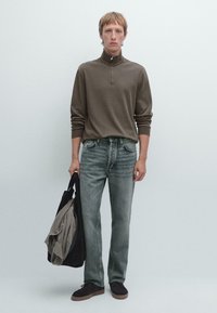 Brauner Reißverschlusspullover, hellgraue Jeans und dunkle Schuhe. Model hält eine schwarze Tasche und ein graues Kleidungsstück. Einfaches, modernes Design, gedämpfte Farben.