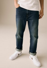 Jeans de mezclilla azul oscuro con corte recto, con puños doblados, combinados con zapatillas blancas con acentos dorados. Camiseta blanca lisa.