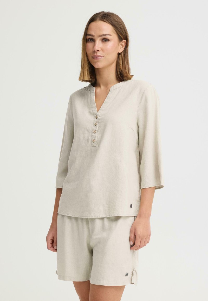 Femme portant un haut ample beige boutonné avec des manches trois-quarts et un short assorti à mi-cuisses sur un fond uni.