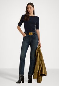 Haut marin à col bateau, jean en denim foncé, bottines noires, et une veste en cuir doré drapée sur un bras. La ceinture présente une boucle dorée.