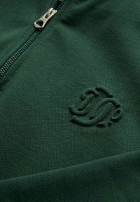 Groene ritsvest van een zachte katoenen mix. Heeft een textuurlogo in de stof, een zilveren rits en een gladde afwerking.