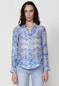 Camicia leggera e trasparente con bottoni, caratterizzata da un motivo geometrico blu e bianco, maniche lunghe con polsini a bottone e colletto.