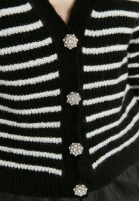 Schwarz-weiße gestreifte Strickjacke mit V-Ausschnitt und fünf runden, mit Juwelen besetzten Knöpfen, die florale Akzente entlang der Vorderseite aufweisen. Strukturierter Strickstoff.