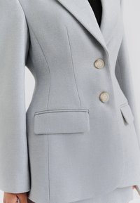 Blazer gris clair sur mesure en tissu texturé. Comprend deux gros boutons, un col cranté et deux poches avant à rabat.