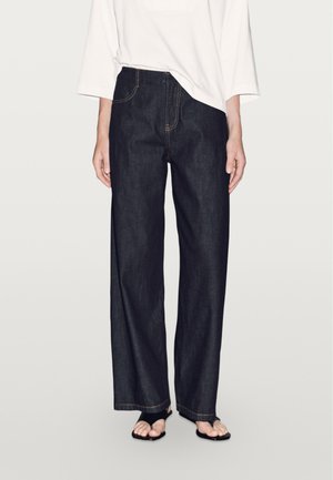 Dark blue wide-leg jeans with visible stitching, a high waist, and front pockets, styled with a loose white top and flat sandals.