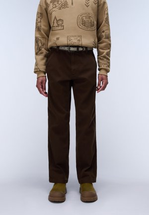 Mand iført brune lige bukser, en beige mønstret sweatshirt, grønne sko og et gråt bælte mod en ensfarvet baggrund.