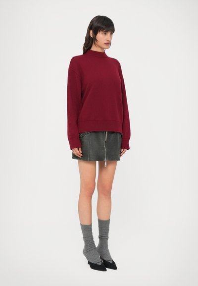 Camisola de malha cor de vinho com punhos canelados, combinada com saia de couro cinza com fecho de correr, meias cinza até ao joelho e sapatos pretos de bico fino.