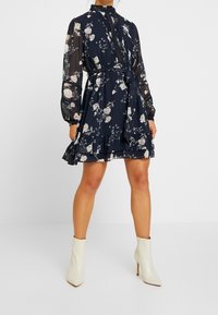 Modell i en marinblå blommig klänning med långa ärmar och volangkant, i kombination med vita ankelstövlar, poserar mot en enkel bakgrund.