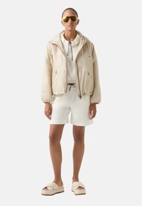 Beige bomber bunda s kapucí, elastickými manžetami, v kombinaci s bílou mikinou a šortkami. Kulaté sluneční brýle a lehká obuv dotvářejí celkový vzhled.