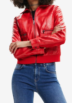 Person mit verschränkten Armen, trägt eine leuchtend rote Lederjacke mit Reißverschlusstaschen und blaue Jeans aus Denim.