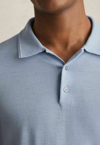 Hellblaues Poloshirt mit weicher Textur, einem Kragen und einer Knopfleiste mit drei Knöpfen. Besitzt dezente Nähte entlang des Halsausschnitts.