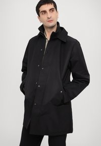 Black waterproof coat with a high collar, snap button closure, side pockets, and a smooth texture. Minimalist design, hip-length fit.