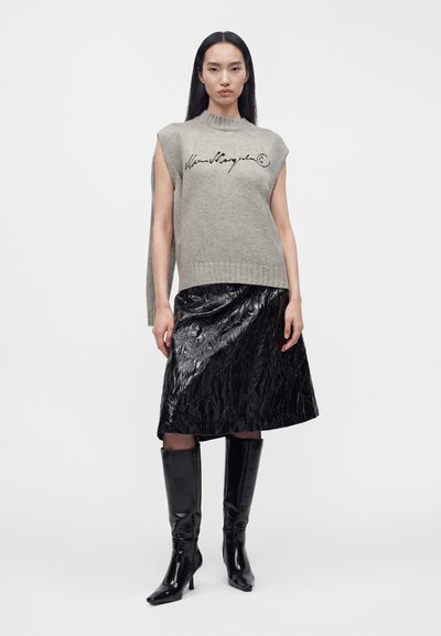 Femme portant un pull sans manches gris avec une écriture noire, une jupe noire brillante longueur genou et des bottes noires montantes, debout devant un fond blanc.