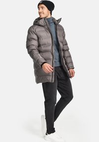 Chaqueta acolchada gris con capucha, con un diseño de acolchado horizontal, cremallera frontal y puños elásticos, llevada sobre un suéter de cuello alto azul y jeans negros.