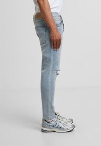 Világoskék, slim fit farmernadrág, enyhe kopással és bézs bőrdísszel a deréknál. Ezüst sportcipővel viselve.