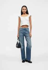 Weißes Cropped-Top mit Rüschensaum, kombiniert mit hellblauen weiten Jeans. Das Model hält eine kleine dunkle Handtasche und trägt schwarze Schuhe.