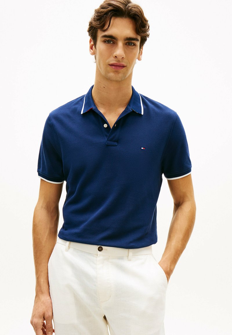 Jeune homme portant un polo bleu marine avec des bordures blanches et un pantalon blanc, se tenant debout avec une main dans la poche devant un fond uni.