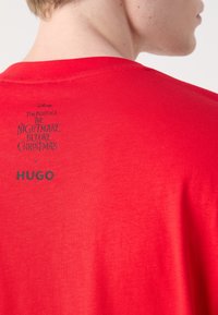 Personne portant un t-shirt rouge avec un texte noir dans le haut du dos indiquant "Tim Burton's The Nightmare Before Christmas x HUGO."