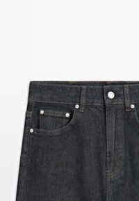 Vita in jeans di denim blu scuro con tasche frontali visibili, passanti per cintura e chiusura con bottone argentato su sfondo bianco.