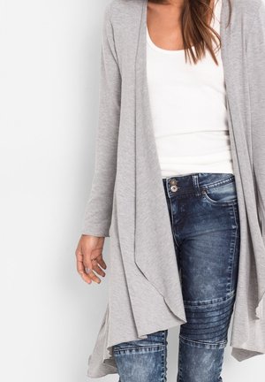 Femme portant un débardeur blanc, un cardigan gris clair drapé et un jean ajusté bleu foncé avec des détails à boutons.