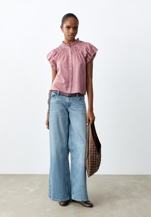 Ženska, oblečena v rožnato bluzo z naborki in široke kavbojke, ki drži rjavo torbico z zakovicami, stoji pred enobarvnim ozadjem.