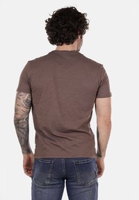 T-shirt marrone a maniche corte con tessuto leggermente texturizzato, scollatura a girocollo e vestibilità rilassata, indossato con jeans in denim blu.
