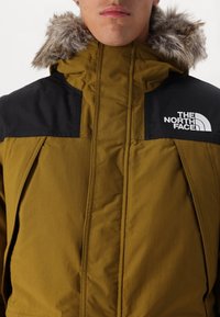 Oliwkowa kurtka z czarnymi panelami na ramionach i kapturem wykończonym sztucznym futrem. Posiada logo na klatce piersiowej, zamek błyskawiczny z przodu oraz boczne kieszenie.