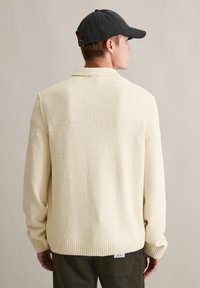 Kremowy, teksturowany sweter z kołnierzykiem i ściągaczami na rękawach, noszony przez osobę w czapce z czarnego materiału i widocznych spodniach z kordurowej tkaniny.