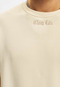 Beige színű pulóver bordázott kerek nyakkivágással és domború "Thug Life" felirattal a mellrészen, puha anyagból, sima textúrával.