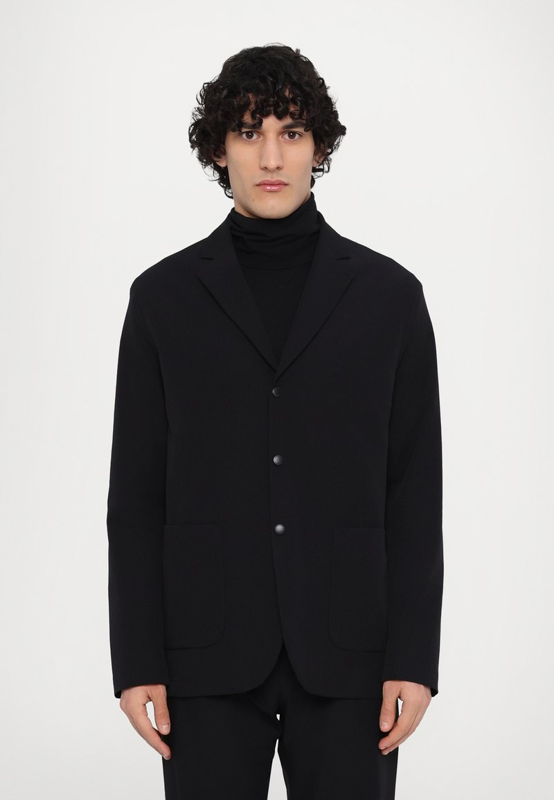 Schwarzes Blazer mit Reverskragen, zwei Fronttaschen und drei silbernen Knöpfen. Getragen über einem schwarzen Rollkragenpullover, glattes Material, taillierte Passform.