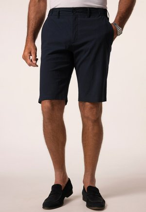 Mand iført mørkeblå, skræddersyede shorts, sorte loafers, hvid skjorte proppet ned, med den ene hånd i lommen og sølvur på håndleddet.