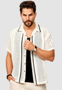 Kurzärmliges Hemd mit Knöpfen aus weißem Netzstoff mit schwarzen vertikalen Streifen. Verfügt über einen Kragen und perlmuttfarbene Knöpfe. Über einem schwarzen Oberteil getragen.