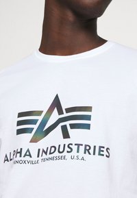 Camiseta de algodón blanca con un logo gráfico multicolor que incluye el texto "ALPHA INDUSTRIES" y detalles de ubicación. Diseño de mangas cortas y cuello redondo.