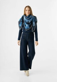 Écharpe à motif floral bleu drapée sur un pull sombre, associée à un jean en denim à taille haute et à jambes larges. Le mannequin se tient devant un fond neutre.