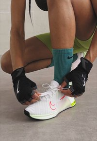 Osoba u tirkiznim Nike čarapama i crnim rukavicama bez prstiju vezuje vezice na bijelim Nike tenisicama s narančastim swooshom.