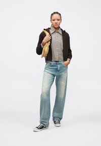 Καφέ φούτερ με φερμουάρ, φορεμένο πάνω από ριγέ πουκάμισο, συνδυασμένο με ανοιχτόχρωμο μπλε τζιν με φθορές και μαύρα αθλητικά παπούτσια. Κρατώντας αξεσουάρ κίτρινου με υφή.